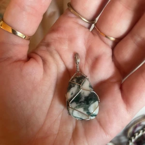 Halsbandspendel med grönvit sten - Unik halsbandspendel med en grönvit, mönstrad sten infattad i silverfärgad metalltråd. Stenen har naturliga mönster och hängs enkelt på en kedja. Perfekt för dig som gillar boho eller vill ha något personligt och snyggt.