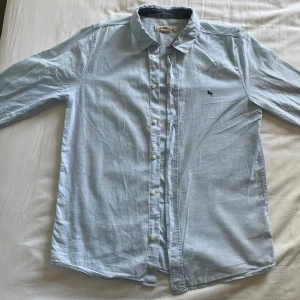 Ljusblå skjorta - Säljer en ljusblå skjorta från L.O.G.G. med klassisk krage och knäppning framtill. Skjortan har lång ärm och en liten broderad get på bröstet. Tillverkad i mjukt linne, perfekt för en clean och stilren look.