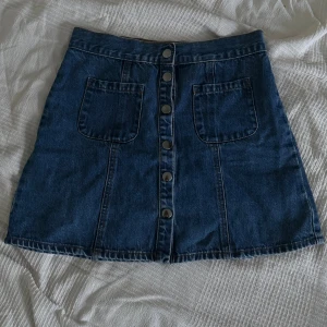 Jeanskjol med knappar från Cooperative - Mörkblå jeanskjol från Cooperative i kort modell med raka linjer. Kjolen har stora fickor framtill och knappar längs hela framsidan.  Mycket fint skick