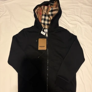 Svart Burberry hoodie - Svart hoodiejacka från Burberry med dragkedja och klassiskt rutigt foder i beige, svart, vitt och rött. Jackan har huva, broderad Burberry-logga på bröstet och stora fickor framtill. Tillverkad i mjuk bomull för en chill och stilren look. Den är tung 