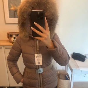 Beige dunjacka Moncler - Säljer en beige dunjacka från Moncler med fluffig äkta päls på huvan, bälte i midjan och dragkedja framtill. Jackan har lång ärm och quiltad design som ger en snygg siluett. Perfekt för kalla dagar och riktigt stylish.