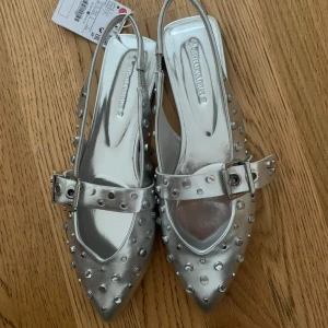 Silvriga ballerinaskor med nitar Stradivarius - Snygga ballerinaskor i metallic silver från Stradivarius. Skorna har spetsig tå, rem med spänne över foten och är täckta med coola runda nitar. Tillverkade i syntetmaterial och med öppen häl för en trendig look.