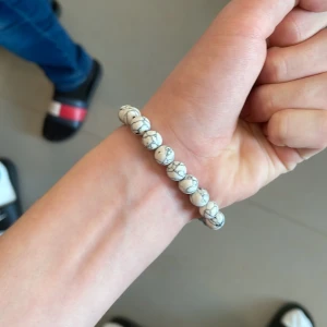 Vitt pärlarmband med marmormönster - Snyggt armband med runda vita pärlor som har ett marmorinspirerat mönster i grått. En av pärlorna är svart med ett coolt sprucket mönster som sticker ut. Perfekt accessoar för dig som gillar stilrena detaljer och unika inslag.