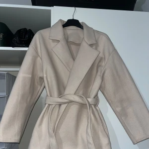 Beige kappa med bälte ( Kappa Matilda)  - Snygg beige kappa med klassisk krage och bälte i midjan. Från Ginatricot. Jackan har en rak passform och är tillverkad i ett mjukt material som känns lyxigt. Perfekt för dig som vill ha en stilren och enkel look under höst och vinter.