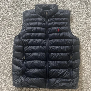 Marinblå  dunväst från Polo Ralph Lauren - Snygg marinblå dunväst från Polo Ralph Lauren med quiltad design och hög krage. Västen har dragkedja framtill, två fickor med dragkedja och den klassiska röda Polo-loggan broderad på bröstet. Perfekt för lager-på-lager och stilren look. Skriv vid minsta fundering!