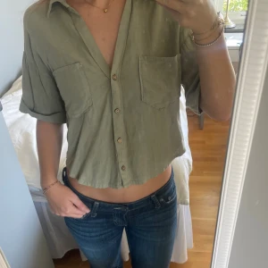 Olivgrön skjorta från Zara - Snygg olivgrön croppad skjorta från Zara i luftigt linnematerial. Skjortan har korta ärmar, två bröstfickor och knappar framtill. 