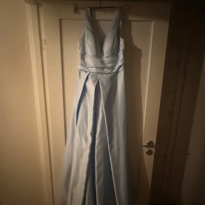 Superfin ljusblå balklänning  - Elegant ljusblå långklänning i satinliknande material med djup v-ringning och breda axelband. Klänningen har markerad midja och ett svepande, utsvängt kjolparti som ger en riktigt drömmig siluett. Perfekt för bal eller student! Endast använd en gång på balen i nian så inga tecken på defekter🤗