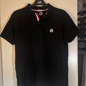 Moncler pikétröja - Snygg svart pikétröja från Moncler med klassisk krage och korta ärmar. Tröjan har Moncler-logga på bröstet och detaljer i rött, vitt och blått vid kragen. Aldrig använd. Perfekt för dig som vill ha en stilren och exklusiv look.