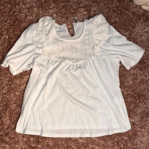 Vit blus med spets från ONLY - Supersöt vit blus från ONLY med korta ärmar och fina spetsdetaljer vid axlarna och bröstet. Blusen har en rund halsringning med liten öppning framtill och ett romantiskt broderat mönster på bröstet. Perfekt för en fräsch och feminin look.