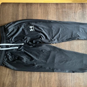 Svarta träningsbyxor Under Armour - Svarta mjukisbyxor från Under Armour med elastisk midja och vita dragsnören. Byxorna har en slim passform och är tillverkade i ett mjukt, syntetiskt material som passar perfekt för träning. Klassisk logga på låret och smala benavslut. Kom med rimliga bud priset är inte fast✌️🙏