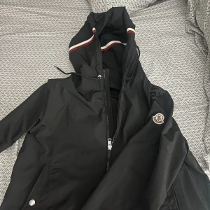 Svart vindjacka från Moncler - Svart vindjacka från Moncler med huva och dragsko. Jackan har dragkedja framtill, fickor med tryckknappar och Moncler-logga på ärmen. Huvan har en snygg randig detalj i rött, vitt och blått. Perfekt för dig som vill ha en stilren och sportig look Den är helt ny jag har tagen o allt på Moncler jag säljer den efter som jag har inte outfit som passar den här vind jackan det är darför jag säljer den jag har knappt använt den inte ens 3 gånger 
