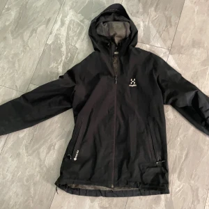 Svart vindjacka från Haglöfs - Svart vindjacka från Haglöfs med huva och meshfoder. Jackan har dragkedja framtill, två fickor med dragkedja och diskret logga på bröstet. Perfekt för blåsiga dagar och outdoor-äventyr. Snygg och funktionell design som funkar till allt.