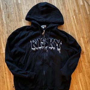 Svart hoodie från Risky med tryck - Svart hoodie från Risky med stort vitt tryck på bröstet i drip-style. Tröjan har huva, dragkedja med coolt R-hänge och två fickor framtill. Perfekt för dig som gillar streetwear och vill sticka ut med en edgy look.