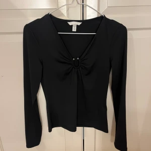 Svart långärmad topp med ringdetalj - Svart långärmad topp från H&M med v-ringning och snygg ringdetalj framtill som ger en cool twist. Toppens material är mjukt och stretchigt, perfekt för att skapa en stilren look. Passformen är normal och toppen är enkel att matcha med olika outfits.