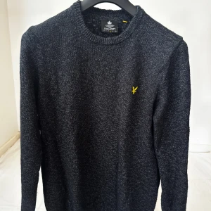 Mörkgrå stickad tröja från Lyle & Scott - Snygg mörkgrå stickad tröja från Lyle & Scott med rund hals och lång ärm. Tröjan har ett litet gult logomärke broderat på bröstet och är tillverkad i mjukt stickat material. Perfekt för dig som gillar stilrena och klassiska plagg. (Storlek L)
