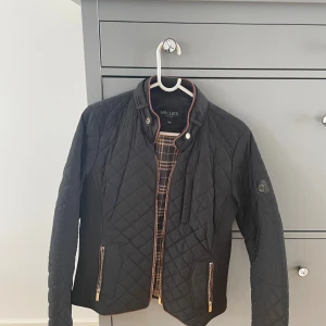 Svart quiltad jacka Hollies Stockholm - Snygg svart quiltad jacka från Hollies Stockholm med guldiga dragkedjor och knappar. Jackan har ett rutigt innerfoder, ståkrage med bruna detaljer och två fickor med dragkedja. Perfekt passform och stilren look för dig som gillar klassisk design.