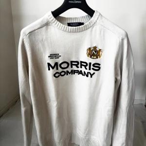 Vit stickad tröja från Morris Company - Vit stickad tröja från Morris Company med broderad logga och text på bröstet. Tröjan har rund halsringning, ribbade muddar och långa ärmar. Perfekt för dig som gillar klassisk och stilren look. Storlek L