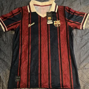 Barça randig fotbollströja S - Snygg FC Barcelona fotbollströja i röd och marinblå med vertikala ränder och klubbmärke på bröstet. Klassisk vit krage och ärmslut med detaljer. Tillverkad i lätt och ventilerande material, perfekt för match eller träning.