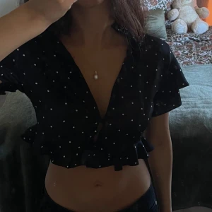 Polka dot topp - Säljer en svart croppad blus med vita prickar och volangkant nertill. Blusen har korta ärmar, djup v-ringning och knappar framtill. Nyskick 
