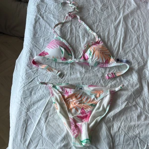 Vit bikini med tropiskt blommönster - Superfin bikini i vitt med tropiskt blommönster i rosa, orange och grönt. Överdelen har trekantskupor och knyt i nacken, medan underdelen har smala sidor. Perfekt för stranden eller poolen i sommar!