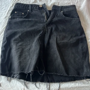 Svart jeanskjol med rå kant - Svart jeanskjol i kort modell med fransig, rå kant nertill. Kjolen har klassiska fem fickor, bälteshällor och dragkedja framtill. Perfekt för dig som gillar en lite edgy och avslappnad stil.