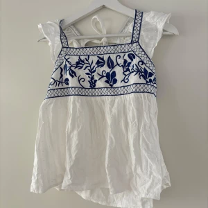 Vit blus med blå broderi från SHEIN - Supersöt vit blus från SHEIN i storlek S med blått broderat blommönster över bysten. Blusen har volangärmar, knytning i ryggen och är gjord i ett lätt bomullstyg. Perfekt för sommarens alla tillfällen!