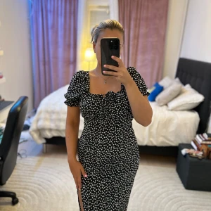 Svart blommig midiklänning från SHEIN - Supersöt svart midiklänning från SHEIN med vita små blommor, puffiga korta ärmar och söt knytning vid bysten. Klänningen har en smickrande slits på sidan och figurnära passform. Perfekt för dig som gillar trendiga och feminina detaljer!