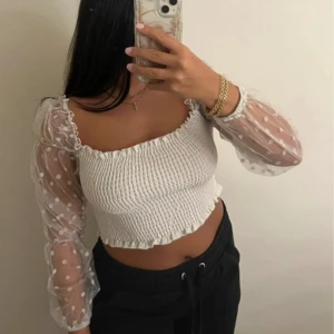 Vit croppad blus med prickiga ärmar - Supersöt vit croppad blus med smockad kropp och transparenta långärmade ärmar i mesh med vita prickar. Toppen har volangkant vid halsringning och nederkant, samt en snygg offshoulder-look. 