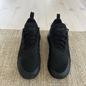 Svarta Nike Air Max 270 sneakers - Snygga helsvarta Nike Air Max 270 sneakers med luftig mesh-ovandel och tydlig Air Max-dämpning i hälen. Skorna har snörning och en sportig vibe, perfekt för dig som gillar streetstyle. Diskret Nike-logga på sidan och dragflik bak för enkel påtagning.
