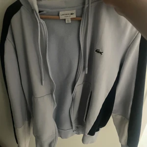 Ljusblå hoodie från Lacoste med dragkedja - Säljer en ljusblå hoodie från Lacoste i klassisk passform. Tröjan har huva med snörning, dragkedja framtill och fickor på sidorna. Mörkblå detaljer längs ärmarna och den ikoniska krokodilloggan på bröstet. Perfekt för chill dagar eller när du vill ha en clean look.