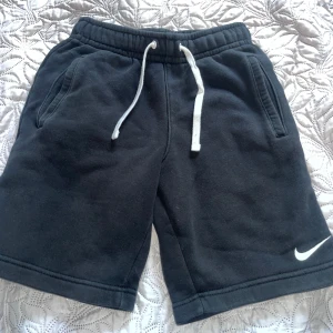 Svarta Nike shorts med snörning S - Svarta mjuka shorts från Nike i storlek S. De har elastisk midja med vit snörning och två sidofickor. Perfekta för chill dagar eller träning. Klassisk Nike-logga broderad i vitt på ena benet. Sköna och stilrena shorts som är lätta att matcha. Passar för 11-12 år