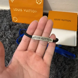 Louis Vuitton svart armband i silver - Stilrent armband från Louis Vuitton med svart band och en rektangulär silverfärgad platta där märkesnamnet är ingraverat. Kommer med originalask och påse. Perfekt accessoar för dig som gillar exklusiva detaljer och enkel design.