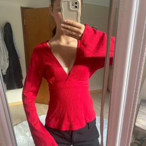 Röd omlottopp från Gina Tricot - Säljer en röd omlottopp från Gina Tricot med långa vida ärmar och djup v-ringning. Toppen har knytband i midjan och är tillverkad i ett lätt, strukturerat tyg. Perfekt för dig som vill ha en färgstark och trendig look. Använd 1 gång 