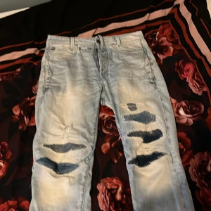 Ljusblå slitna jeans G-Star RAW 28/32 - Ljusblå jeans från G-Star RAW, modell D-Staq, med snygga slitningar och hål på benen för en trendig look. Jeansen har knäppning med knappar, fem fickor och är tillverkade i klassisk denim.
