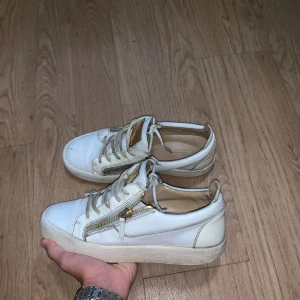 Vita sneakers från Giuseppe Zanotti - Snygga vita sneakers från Giuseppe Zanotti med guldfärgade metalldetaljer och dragkedjor på sidorna. Skorna har snörning framtill, platt sula och är tillverkade i skinn. Perfekta för dig som gillar exklusiva och trendiga sneakers med unika detaljer.