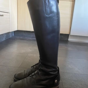 Svarta höga ridstövlar i skinn - Snygga svarta ridstövlar i äkta skinn. Klassisk modell med snörning framtill och rund tå. Stövlarna är höga och har en stilren look med diskret logga på utsidan. Perfekta för dig som vill ha en clean och tidlös stil i stallet.