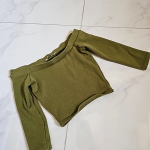Olivgrön offshoulder topp från Zara - Säljer en olivgrön offshoulder topp från Zara i ribbad bomull. Toppen har långa ärmar och en croppad passform som ger en snygg siluett. Perfekt att styla med höga jeans eller kjol för en trendig look.