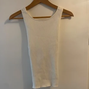 Vit ribbad linnetopp med öppen rygg - Säljer en vit ribbad linnetopp med breda axelband och snyggt korsad öppen rygg. Toppen är ärmlös och har en tight passform som framhäver figuren. Materialet är mjukt och stretchigt.