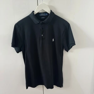 Svart pikétröja Polo Ralph Lauren M - Svart pikétröja från Polo Ralph Lauren i slim fit med klassisk krage och knappar framtill. Broderad logga på bröstet och tillverkad i stretchig bomullsmix som ger en skön passform. Perfekt för en clean och stilren look. Skriv vid frågor!