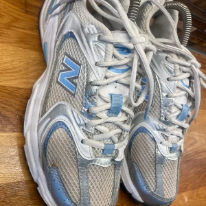 New Balance 530 sneakers blå/vit - Snygga New Balance 530 sneakers i vitt och ljusblått med meshpaneler och silvriga detaljer. Skorna har snörning, platt sula och rund tå. Perfekta för dig som gillar sportig och trendig stil. Ikonisk N-logga på sidan och bekväm dämpning med Abzorb-sula.