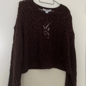Brun stickad tröja med hålmönster - Mysig brun stickad tröja med lång ärm och snyggt hålmönster. Tröjan har en något croppad passform och rund halsringning. Perfekt för lager-på-lager och ger en chill vibe till din outfit.