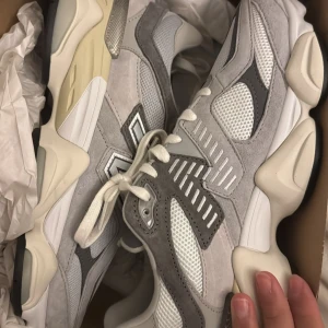 New Balance grå 9060 - Säljer ett par feta New Balance sneakers i grått och vitt med chunky sula och coola röda detaljer undertill. Ovandelen är i mesh och mocka med stora N-loggan på sidan. Skorna har snörning och en riktigt modern vibe, perfekta för dig som gillar streetstyle.