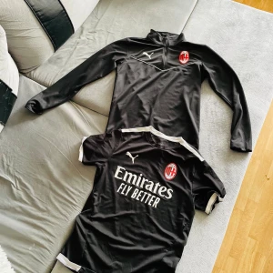 AC Milan svart fotbollströja Puma - AC Milan svart fotbollströja från Puma med vit logga och klubbmärke på bröstet. Tröjan har korta ärmar och vita detaljer vid axlar och ärmslut. Materialet är lätt och andas, perfekt för träning eller match. Snygg design med trycket 'Emirates Fly Better' på framsidan.