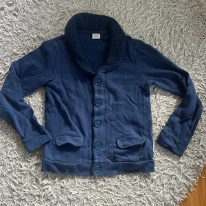 Cardigan - Snygg mörkblå kofta från Polarn O. Pyret med grovstickad krage och stora knappar framtill. Koftan har två fickor och långa ärmar, perfekt för dig som gillar klassisk stil med en twist. Materialet är mjukt och känns skönt mot huden. Passar bra nu till hösten.