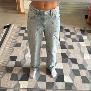 Ljusblå jeans med broderade blommor - Säljer ett par högmidjade ljusblå jeans med broderade små blommor i grönt och orange. Byxorna har rak passform, klassisk femficksdesign och normal midja. Perfekta för dig som vill ha ett par unika och snygga jeans med lite extra detaljer.