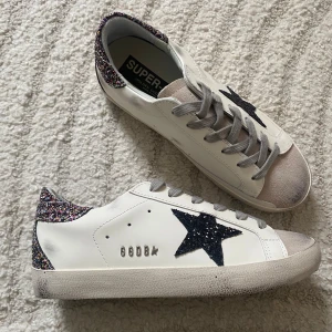 Golden Goose Superstar sneakers med glitter - Golden Goose Superstar sneakers i vitt skinn med grå mockadetaljer på tån, grå snörning och svart glittrig stjärna på sidan. Hälkappan är täckt av färgglatt glitter som ger extra edge. Klassisk rund tå och platt sula. GGDB-logga i silver på sidan. Pris går att diskutera!💓
