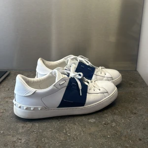 Valentino Garavani sneakers vit/blå - Snygga sneakers från Valentino Garavani i vitt skinn med en bred blå lackdetalj över mitten. Klassisk rund tå, vita snören och nitar baktill på sulan. 