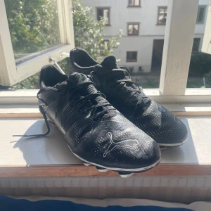 Svarta fotbollsskor från Puma - Svarta fotbollsskor från Puma med vita detaljer och logga på tån. Skorna har snörning och mönstrad ovandel i syntetmaterial. Sulan har fasta dobbar för bra grepp på planen. Perfekta för dig som vill ha stilrena och sportiga skor till matchen.