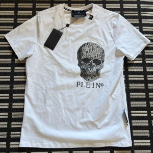 Vit Philip Plein Tshirt Helt Ny - Snygg vit t-shirt från Philipp Plein med ett stort döskalletryck på bröstet där märkesnamnet är integrerat i designen. Klassisk rund hals och korta ärmar. Tillverkad i mjuk bomull som känns skön mot huden. Perfekt för dig som gillar edgy streetstyle.
