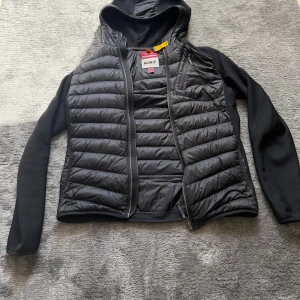 Svart hybridjacka från Parajumpers - Svart hybridjacka från Parajumpers med quiltad framsida och rygg, huva och släta ärmar i stretchigt material. Jackan har dragkedja, bröstficka med dragkedja och flera innerfickor. Perfekt för lager-på-lager och snygg streetstil.